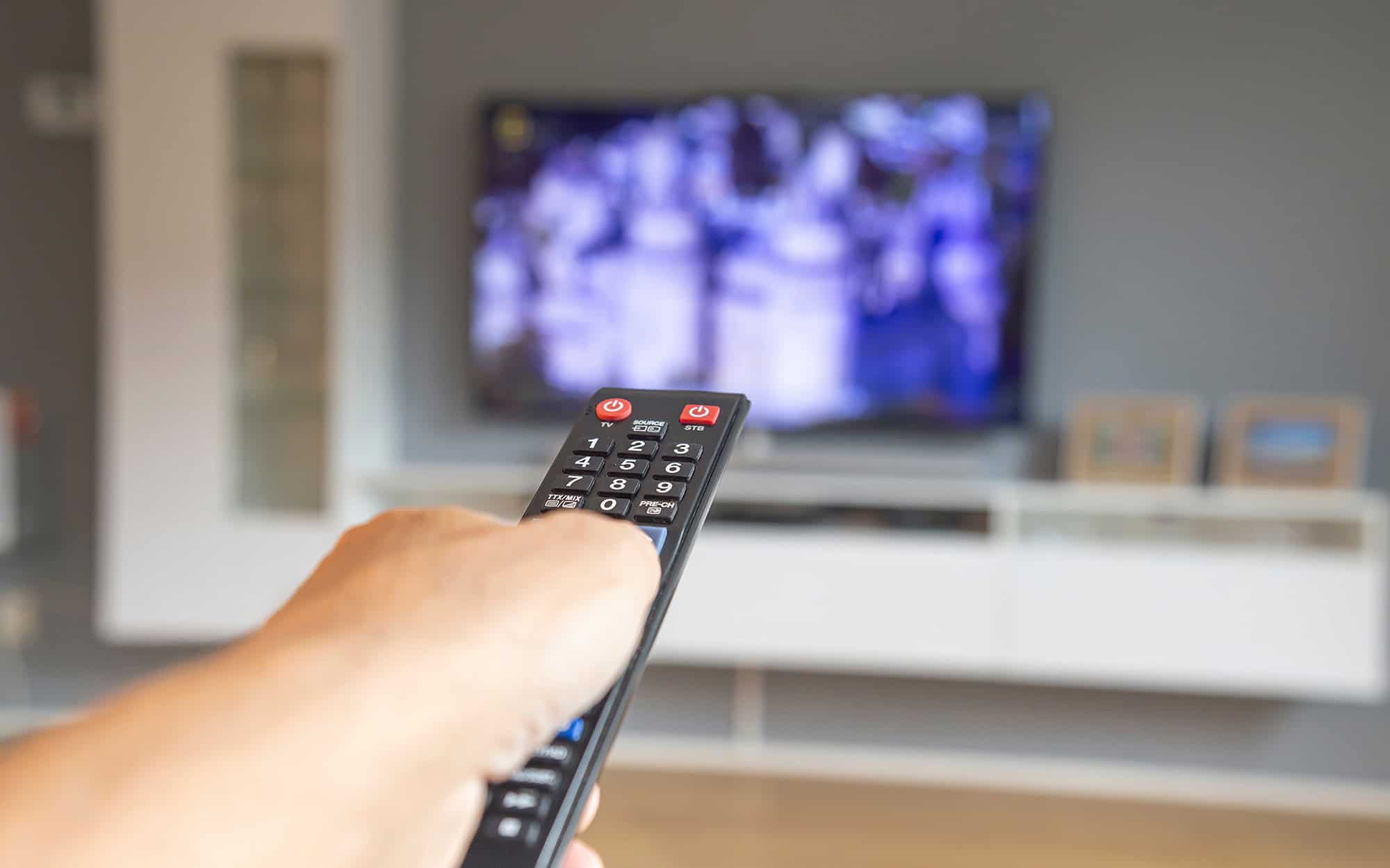 Closeup to a male hand holding a black TV controler with a blurred flat tv at background